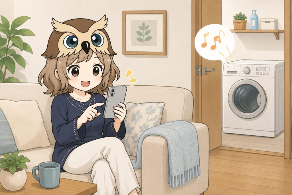 フクロウ帽子の女の子が、洗濯機が止まったことに気づかずにスマホを見ている様子。洗濯機が稼働終了音を鳴らしているが気づいていない。